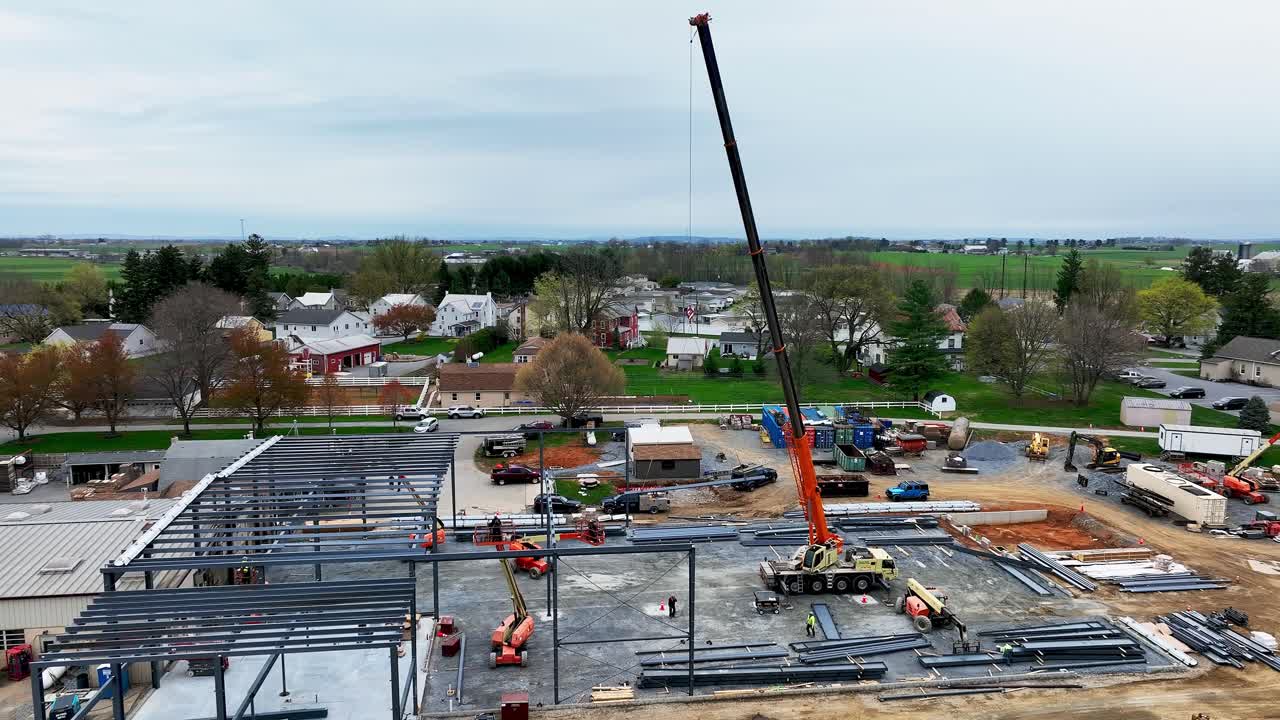 Workers are busy at a construction site in a small town. They are setting up steel beams and preparing the ground for a new building. Nearby homes and farmland are visible under a cloudy sky