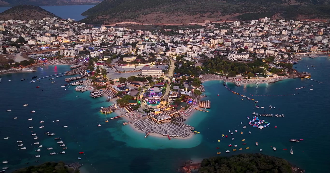 Toàn cảnh bãi biển Ksamil và thị trấn ven biển từ trên cao lúc hoàng hôn, Albania. Cảnh biển buổi tối tuyệt đẹp với ánh đèn neon, các khu nghỉ dưỡng du lịch, những chiếc thuyền trên làn nước màu ngọc lam, Biển Adriatic Ionian.