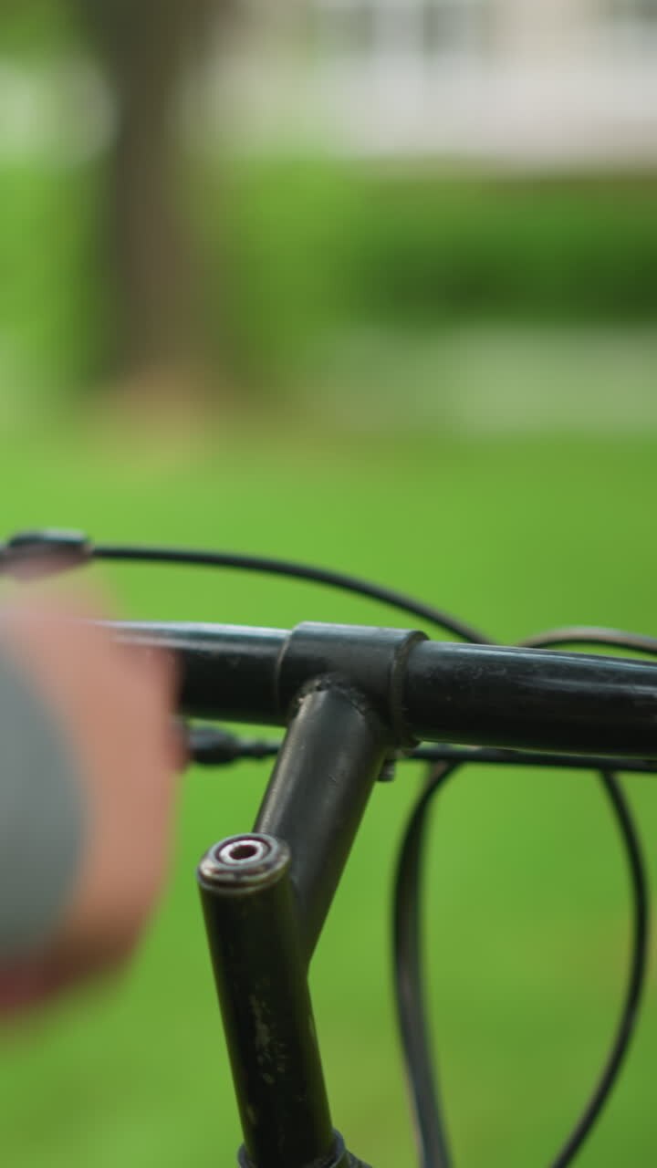 Close-up of parked bicycle with partial view of hand in gray clothing holding the handlebar of bicycle, observing the bike, the background is blurred, with a lush green park