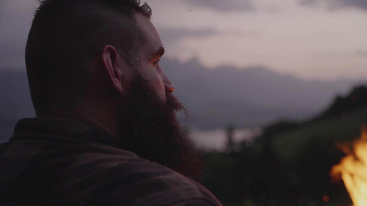 A man sits beside a glowing campfire, gazing toward the mountains during sunset. Peaceful outdoor moment symbolizing solitude, reflection, and connection with nature