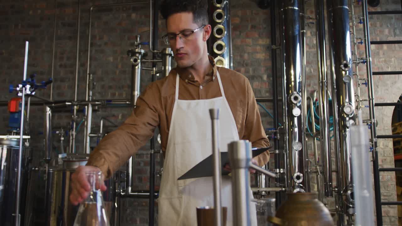 Caucasian man working at distillery checking gin product in flask and holding clipboard