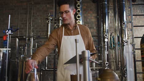 Caucasian-man-working-at-distillery-checking-gin-product-in-flask-and-holding-clipboard