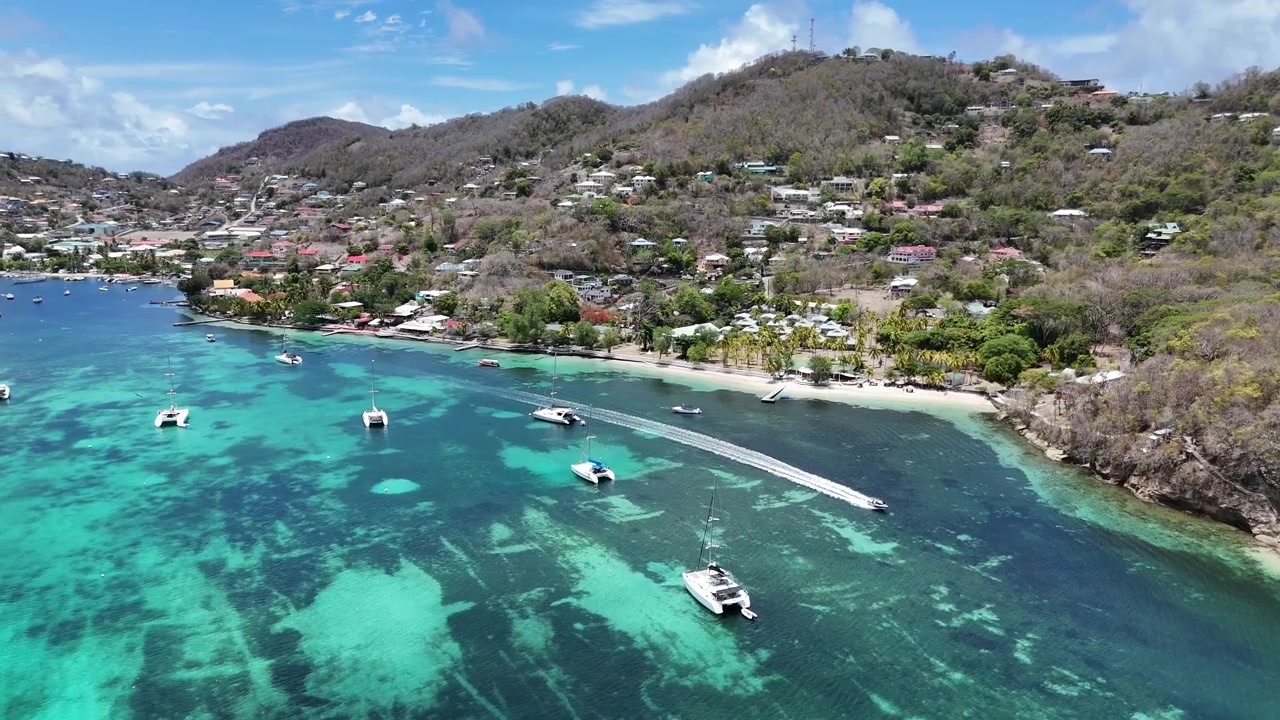 Vista aerea bassa con drone di barche ancorate nelle vivaci acque turchesi cristalline della barriera corallina lungo le lussureggianti colline costiere dell'isola di Bequia, Saint Vincent e Grenadine