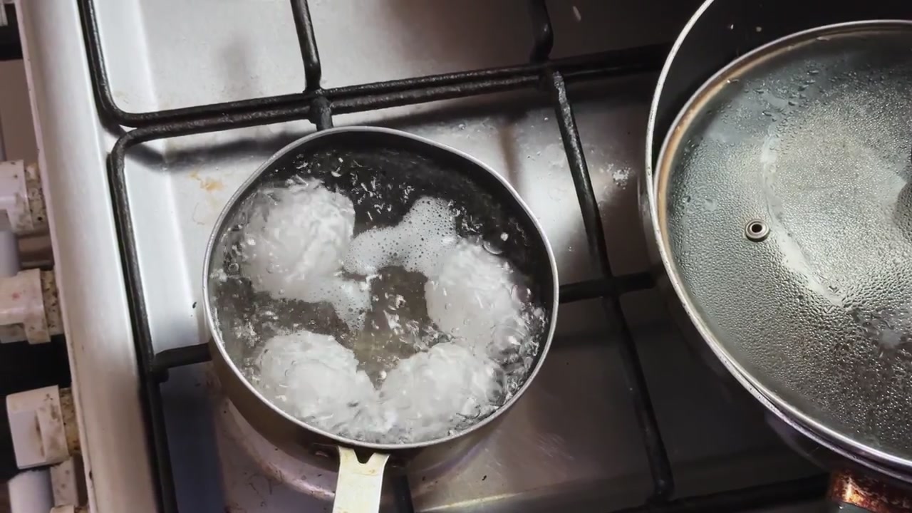 Top-down view of white eggs boiling in hot bubbling water inside a small pot on a kitchen gas stove during the day. This close-up shot highlights a healthy breakfast preparation process.