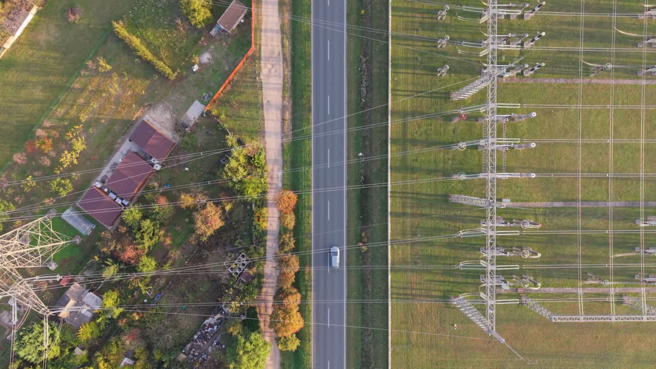 A drone aerial video showing cars driving on a country road surrounded by green fields and power lines. Electricity pylons and cables stretch across the landscape