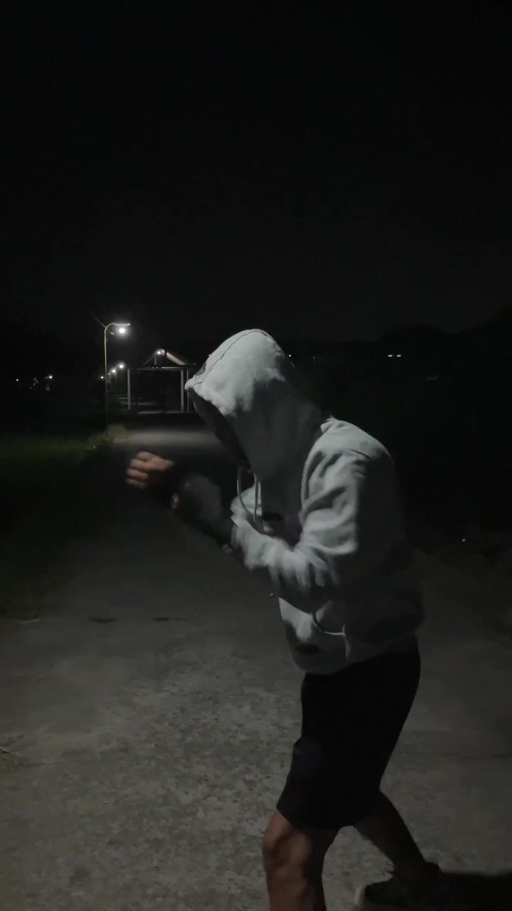 Vertical 4K slow motion rotating camera medium shot from behind of a male athlete in hoodie doing shadow boxing training under street light lamp posts at dark night Sport fitness broll