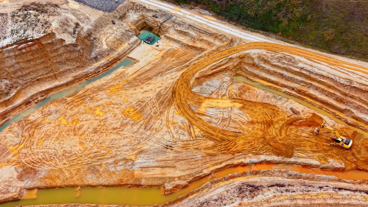 Drone view of layered sand quarry walls with teal water channel, Bulgaria. Oblique perspective showing orange and beige sediment layers, tire tracks, flooded trench and one vehicle on plateau.