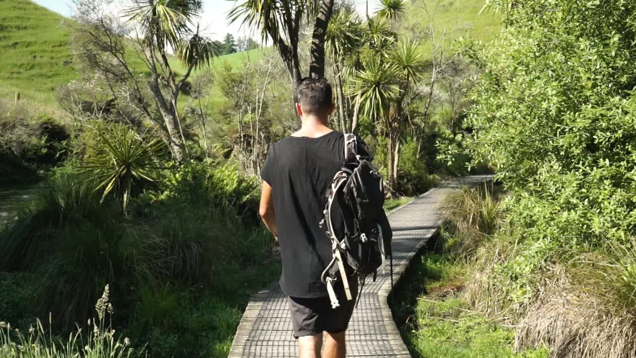 Caucasian male tourist from behind walks along Putaruru Blue Sprinng river surrounded by native lush New Zealand forrest