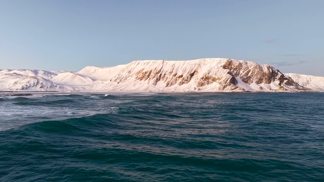 Aerial drone gliding close and flying over dark blue and green wavy ocean sea water with mountains in the background under sunshine in Norway during winter time. Footage is flying towards the beach.