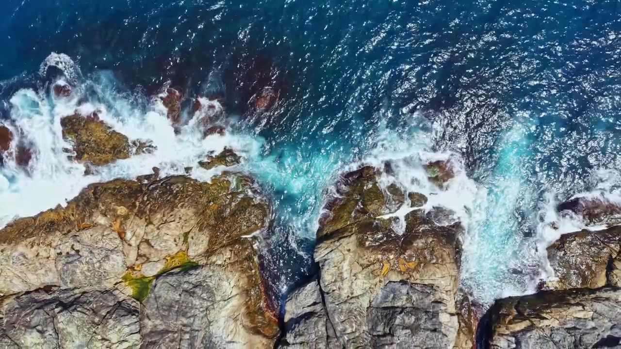 Wellen branden an felsiger Küste mit blauem Wasser in einem Küstengebiet unter der Mittagssonne.