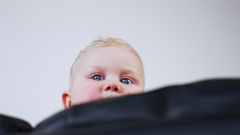 lovely blond baby peeping out from behind the sofa. serious grey eyed infant kid portrait. low angle view.