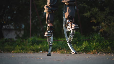 lower body of person jumping while wearing loose grayish-brown cargo pants tucked into custom-made metallic silver spring stilts. captured against blurred green park background