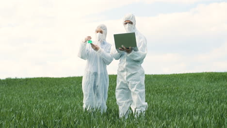 caucasian researchers in protective suit holding a test tube and using laptop while doing pest control in the green field