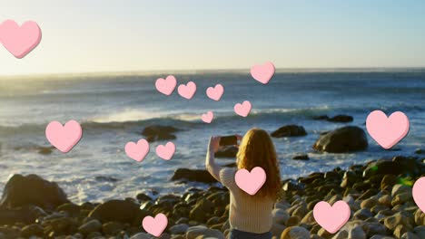 Animation-of-hearts-over-back-view-of-caucasian-woman-taking-photo-on-beach