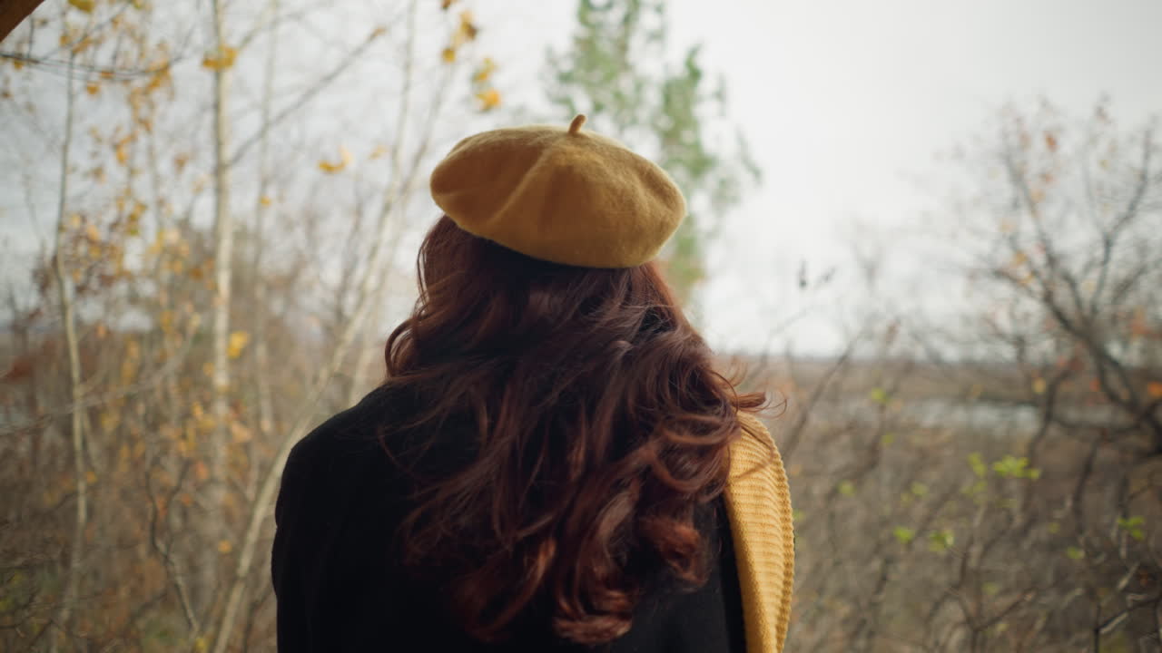 Vista trasera de una joven con boina amarilla apoyando sus manos en una barandilla de madera, contemplando el bosque mientras las hojas de otoño se mecen con el viento.