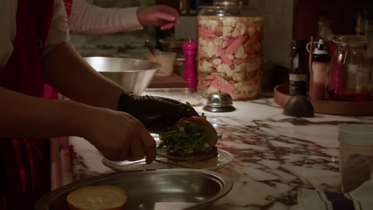 A professional chef wearing a red apron carefully places the top bun on a gourmet slider topped with fresh arugula in a high-end restaurant kitchen