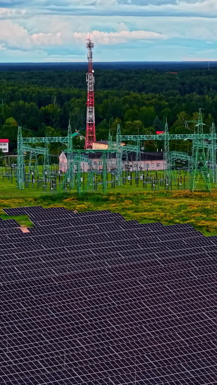 Solar panels are installed on a sunny field in front of an electrical station. A forest lies in the background