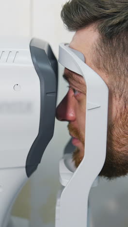 patient man looks at device in ophthalmology clinic. profile of patient's face looking into apparatus for eyes diagnostic in clinic. close-up. vertical video