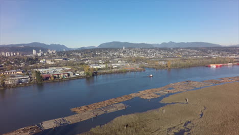 Antena-De-Madera-Flotante-Y-Remolcador-En-El-Río-Con-Una-Ciudad-En-El-Fondo