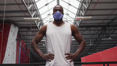 fit african american man wearing face mask standing with hands on his hips in the gym