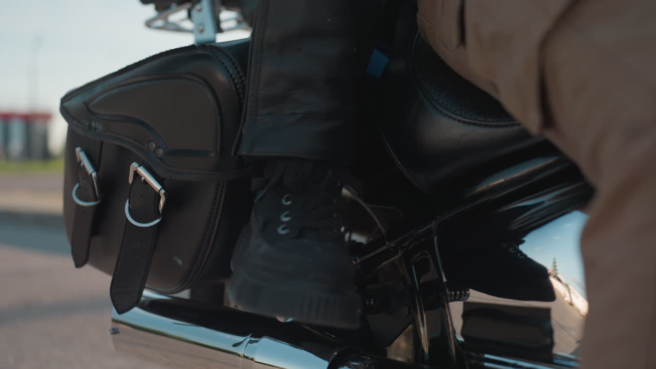 Close up of motorcycle lower section showing rider seated on chrome engine bike with tan pants and black boots while passenger begins climbing on, captured with urban background