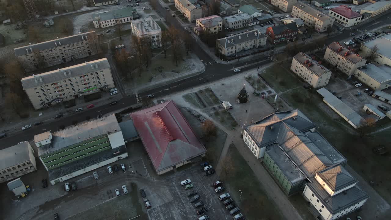 Aerial photo of Balvi city bathed in warm sunset light, featuring urban buildings, parking lots, and a snow-dusted landscape. A peaceful winter scene.