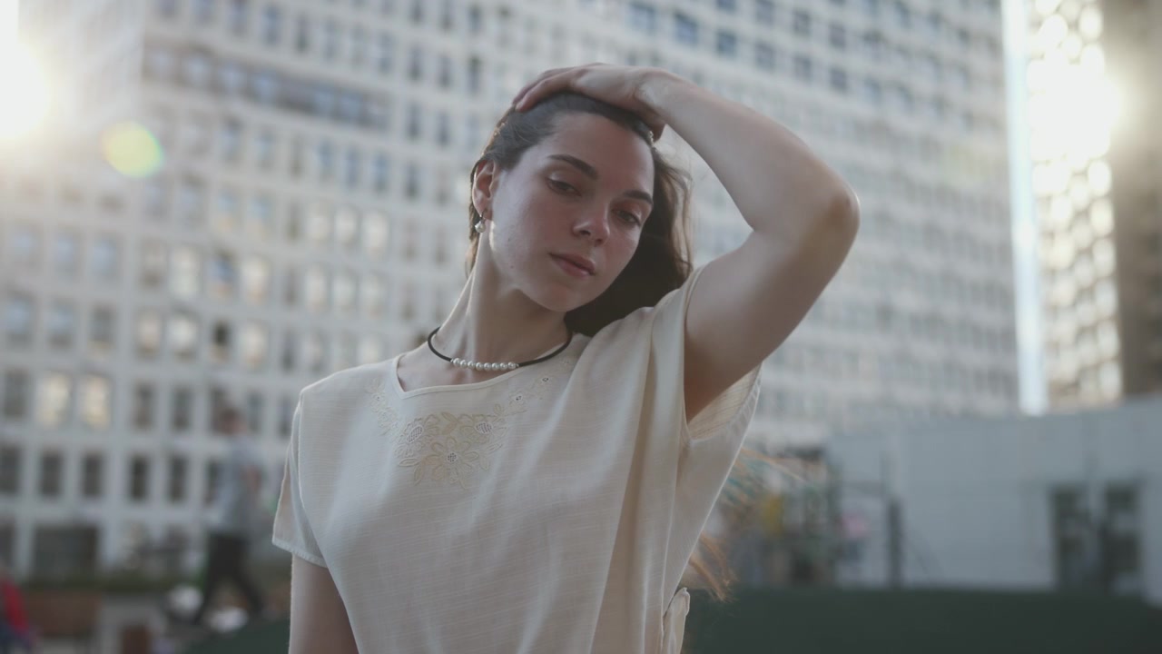 Donna in piedi in una piazza urbana accanto a alti grattacieli, che gira la testa e si accarezza i capelli mentre una leggera brezza solleva le ciocche, retroilluminazione calda, espressione calma, atmosfera di vita moderna in città