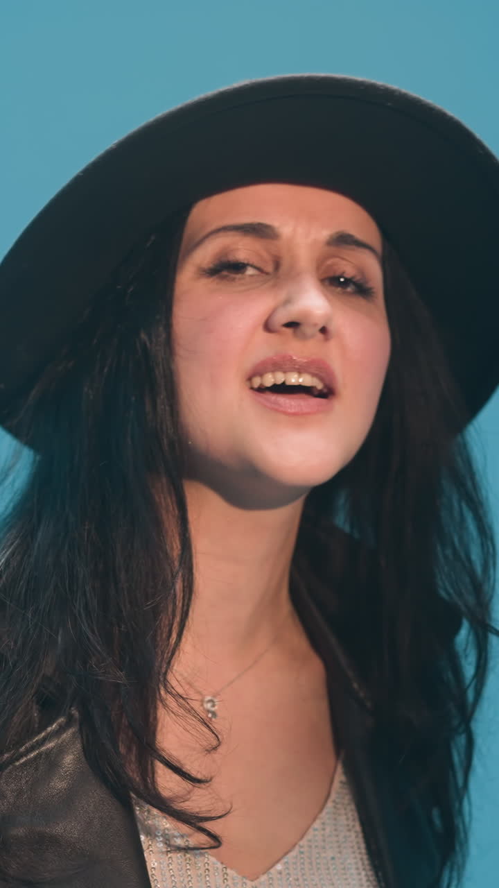 primer plano de una dama con un sombrero negro y una chaqueta de cuero negra, cantando apasionadamente con la mano levantada. un tecladista borroso es visible en el fondo contra un telón de fondo azul