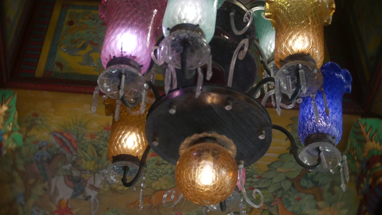 Lampadario con paralumi in vetro colorato che brillano in alto, più lampadine che illuminano tonalità vivaci di rosa, blu e ambra. Opera d'arte decorativa sul soffitto visibile sopra, creando un ambiente interno ricco e ornato.