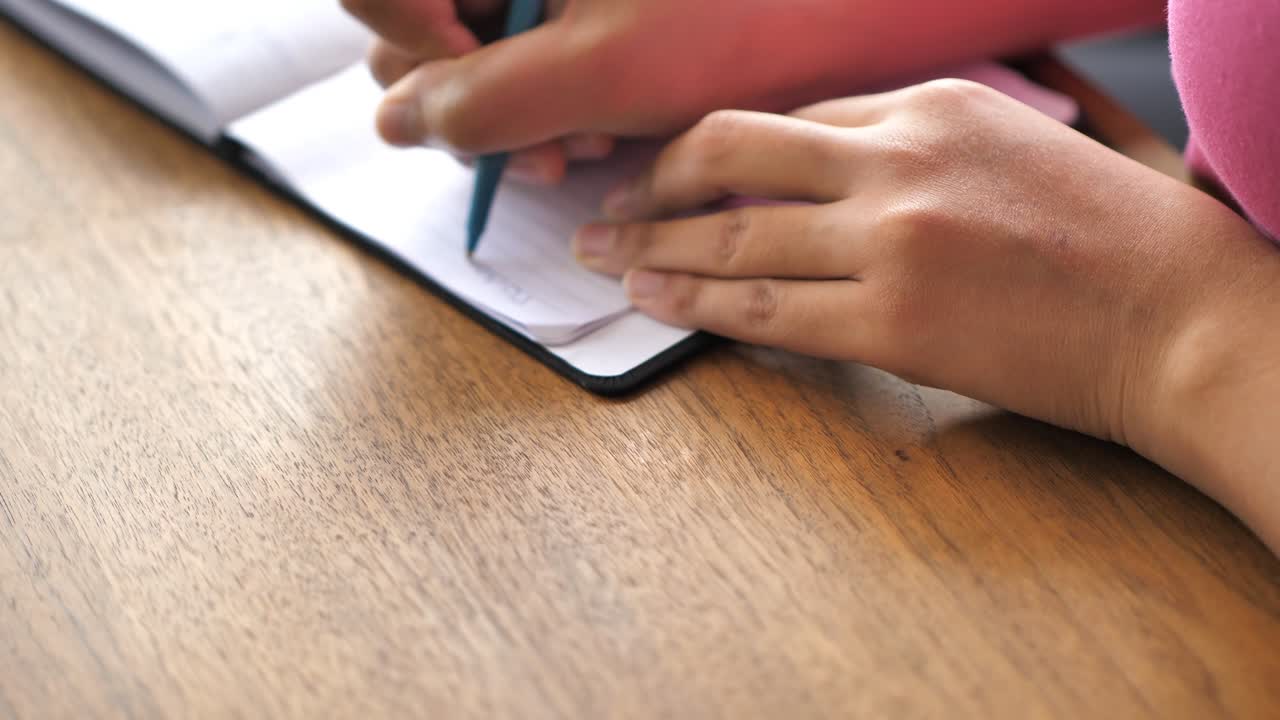 A closeup view of a black mixed race African American woman wearing a pink shirt sitting at a wood dining table and writing a note in her paper notepad with a pen with her hands and chest in frame.