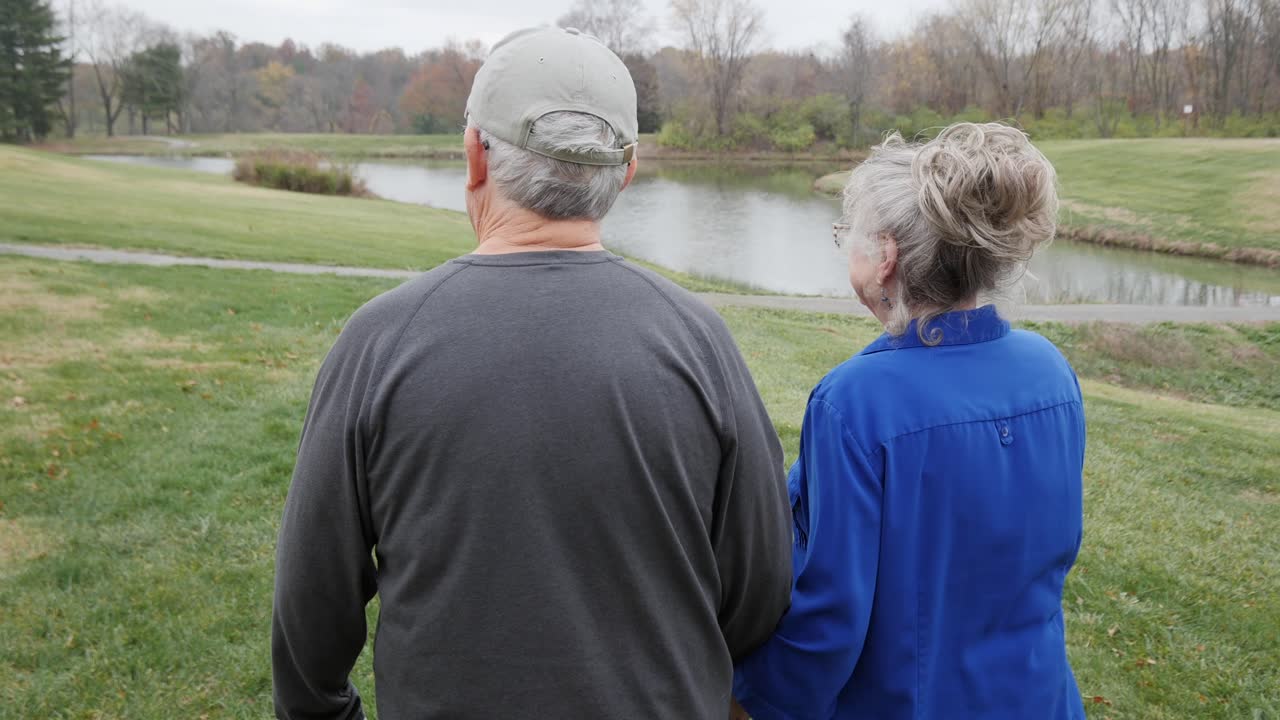 Following an elderly couple walking toward pond in the park on an autumn day