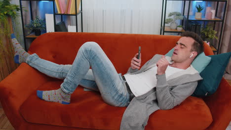 happy relaxed man in wireless earphones dancing, listening favorite music, lying on couch at home