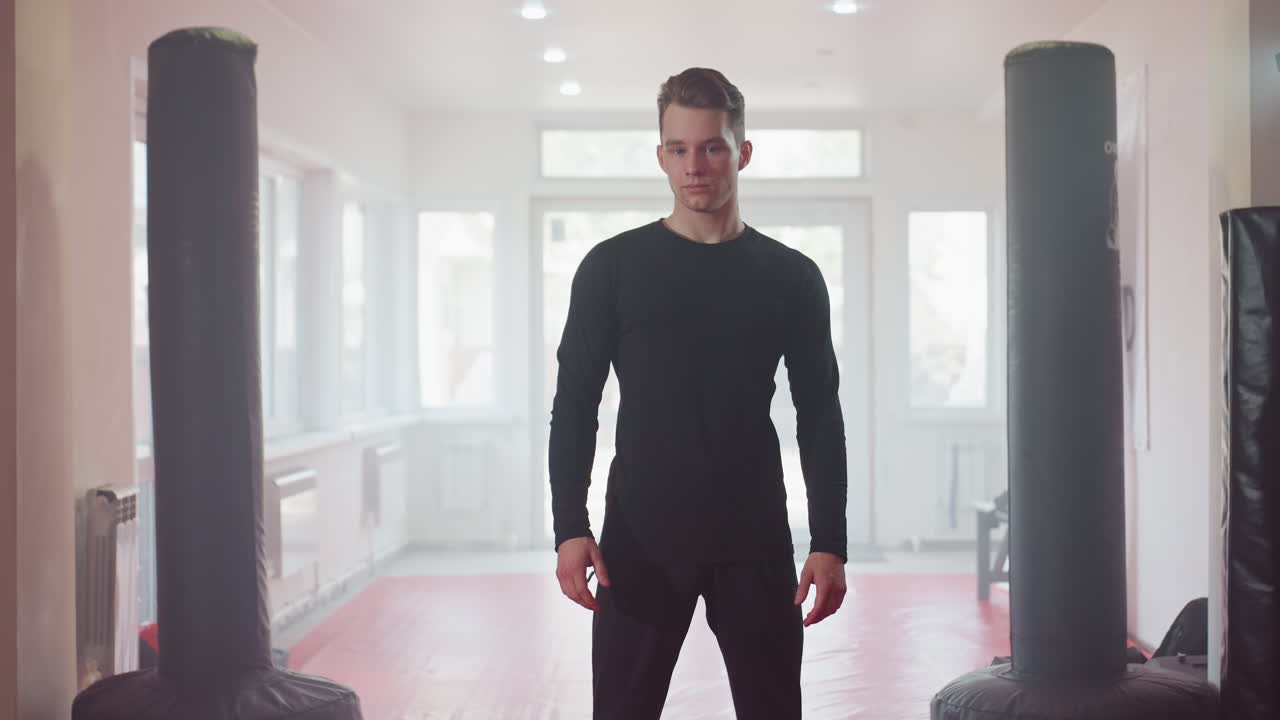 Gentleman in black long sleeve shirt stands in bright gym environment with natural light, gazing confidently to side, showcasing sharp features, hairstyle, and tattooed neck while embodying modern
