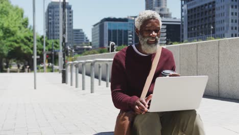 Afroamerikanischer-älterer-Mann-Hält-Kaffeetasse-Mit-Laptop-In-Der-Hand,-Während-Er-Auf-Der-Treppe-Eines-Unternehmens-Sitzt