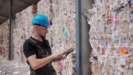 Worker-at-paper-recycling-plant-counts-pressed-paper-bales,-make-notes-on-tablet