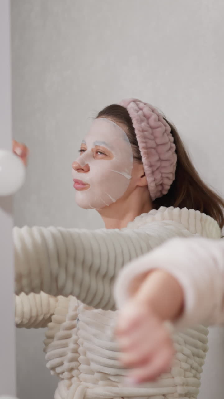Lady takes care of facial skin performing dance moves. Woman puts fabric moisturizing mask on face enjoying day off listening music slow motion