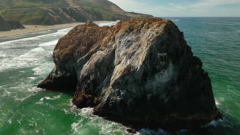 Aerial-view-of-a-large-rock-sitting-in-the-water-of-California's-coast,-slowly-eroding