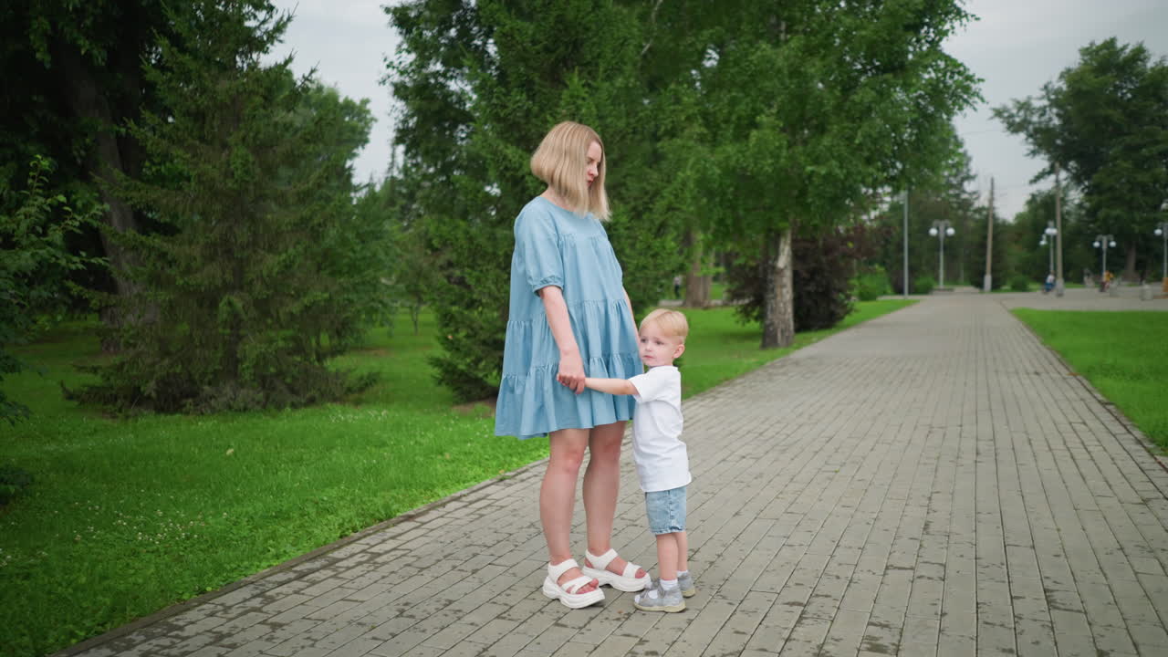 A tender moment between a mother and child holding hands while standing on a paved walkway surrounded by a lush green park
