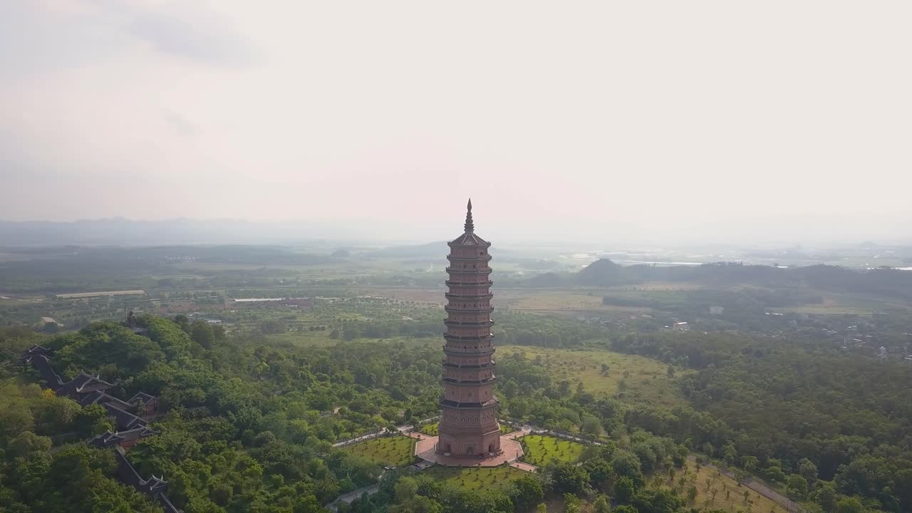 Vista aérea con dron de la alta torre de la Pagoda Bai Dinh rodeada de un paisaje de bosque verde y el campo distante, creando un sereno escenario de referencia budista en Ninh Binh, Vietnam.