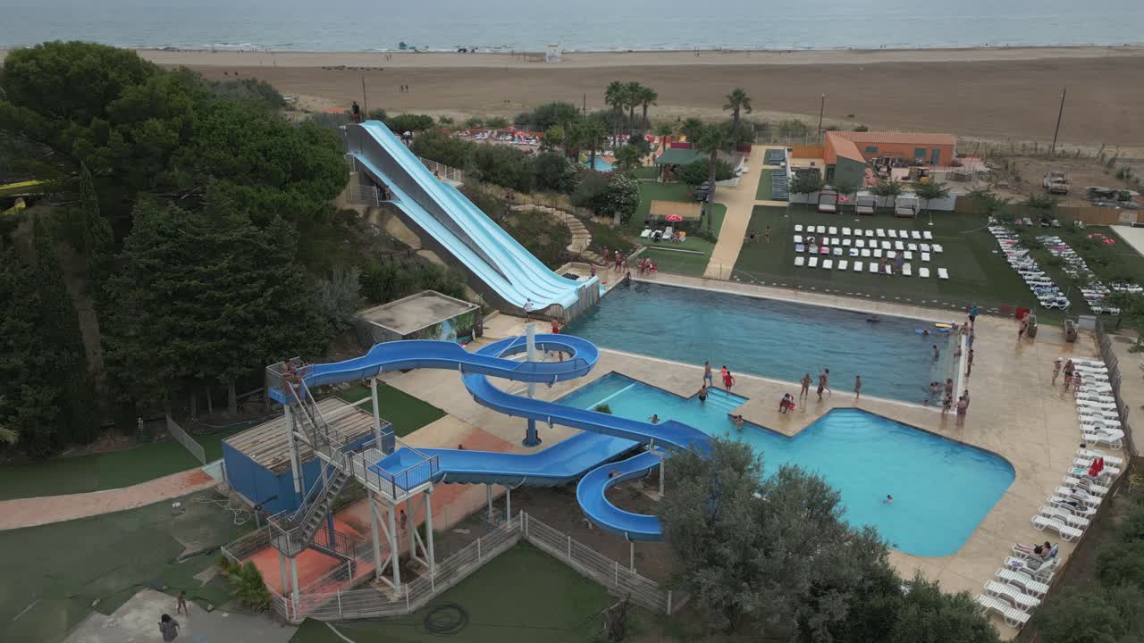 Families enjoy a sunny day at a coastal water park featuring multiple swimming pools and exciting water slides. The beach is just beyond the park, perfect for relaxing