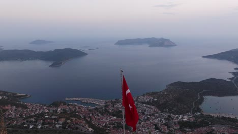 turkish flag on a hilltop overlooking kas at dusk, with a serene coastal backdrop of town along idyllic coast, aerial dolly tilt down