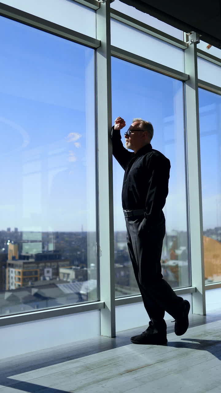 Thoughtful adult businessman comes up to a panoramic window. Man looks a sunny cityscape from upper floor office. Vertical video.