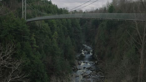 Narrow-Rocky-River-Between-Lush-Trees-Under-Suspension-Bridge-At-Coaticook-In-Eastern-Townships,-Quebec-Canada,-Tilt-Up-Shot