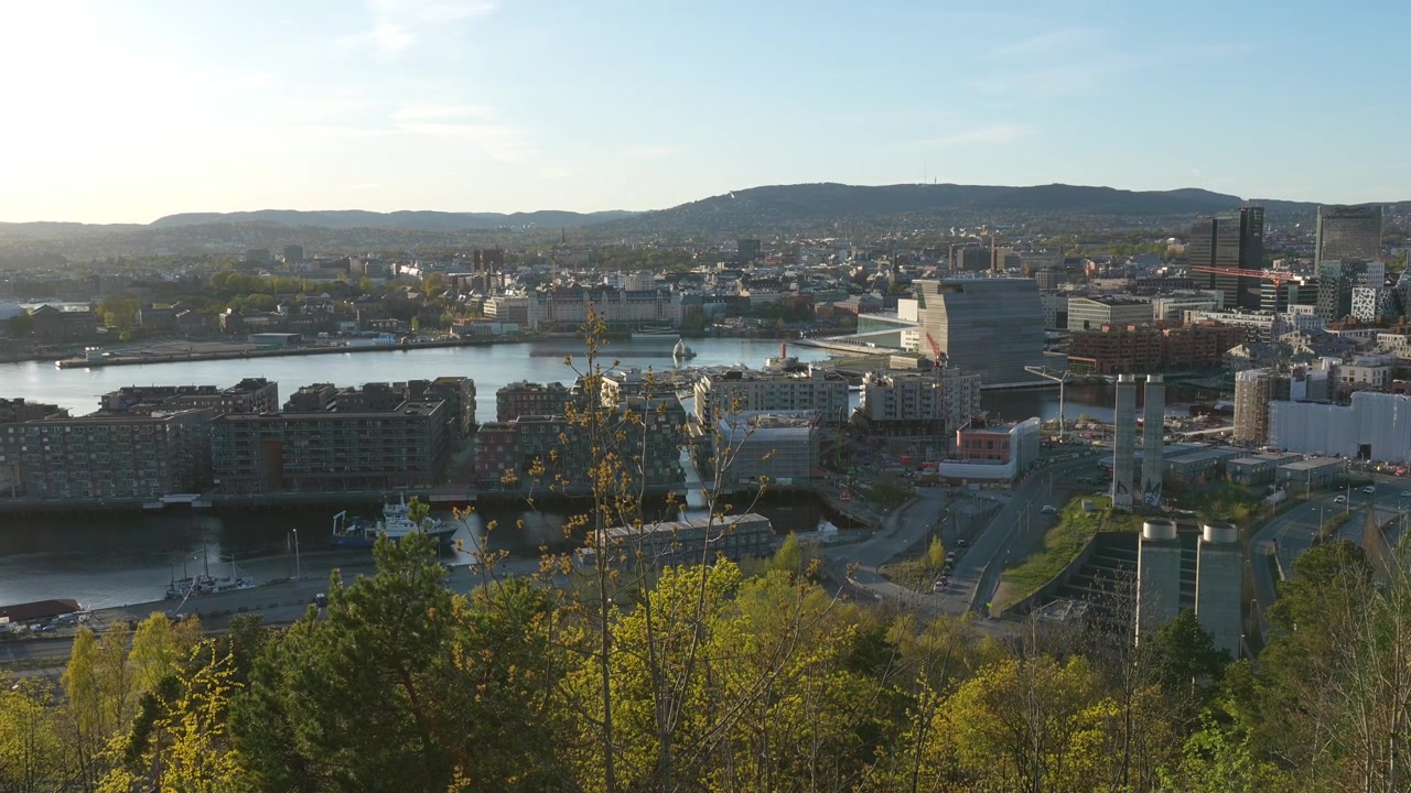 Weitläufige Panoramaaufnahme der Stadt Oslo zur goldenen Stunde vom Vaterland Park aus, die moderne Architektur, den Hafen, üppiges Vordergrundlaub und ruhiges Abendlicht zeigt. Keine Personen sichtbar.