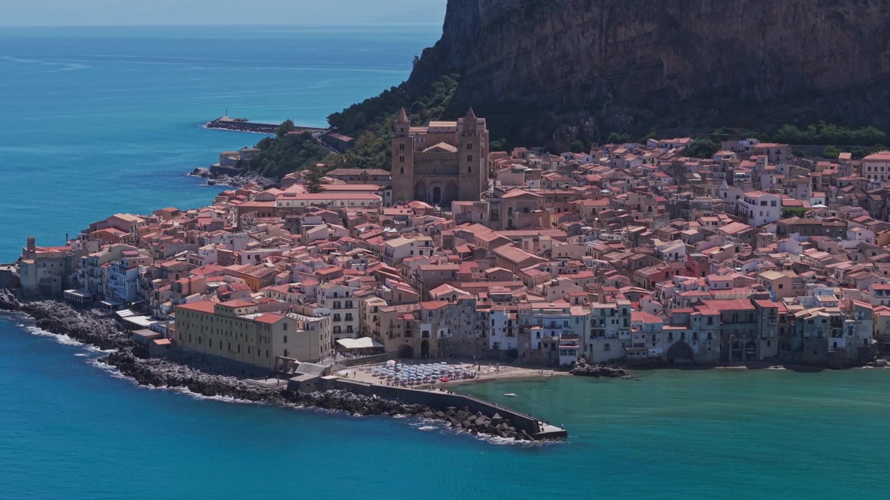 Una vista aerea di una storica città costiera mediterranea, caratterizzata da una cattedrale imponente, edifici fittamente raggruppati e acque turchesi cristalline.