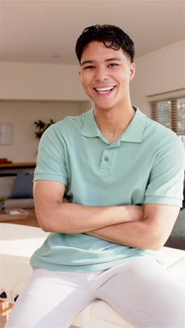 vertical video: smiling young man in casual attire crossing arms while sitting indoors