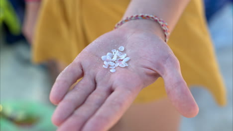 cerca de la mano de una mujer caucásica sosteniendo lindas pequeñas conchas blancas en la playa con una falda amarilla y una pulsera