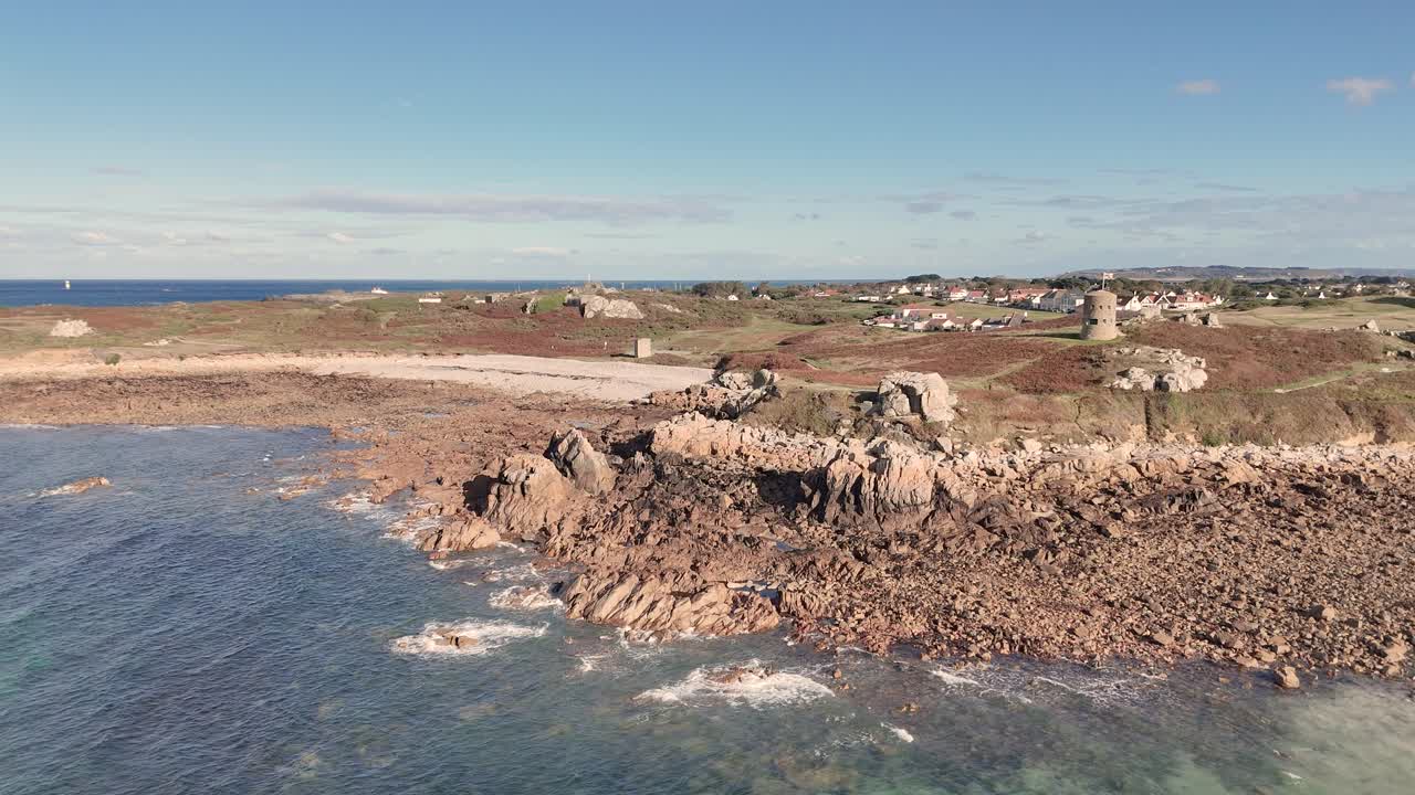 Lancresse Bay Guernsey flight towards shoreline of beach with rocky outcrops on sunny day with calm lapping waves showing Martello Tower and Lancresse Common