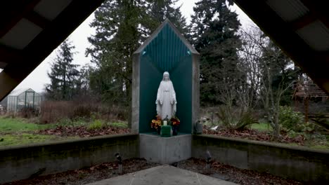 Estatua-De-La-Virgen-María-En-Un-Altar-Fuera-De-Una-Pequeña-Capilla-De-Madera-En-El-Exuberante-Parque-Sin-Gente---Tiro-De-Retroceso-De-Drones