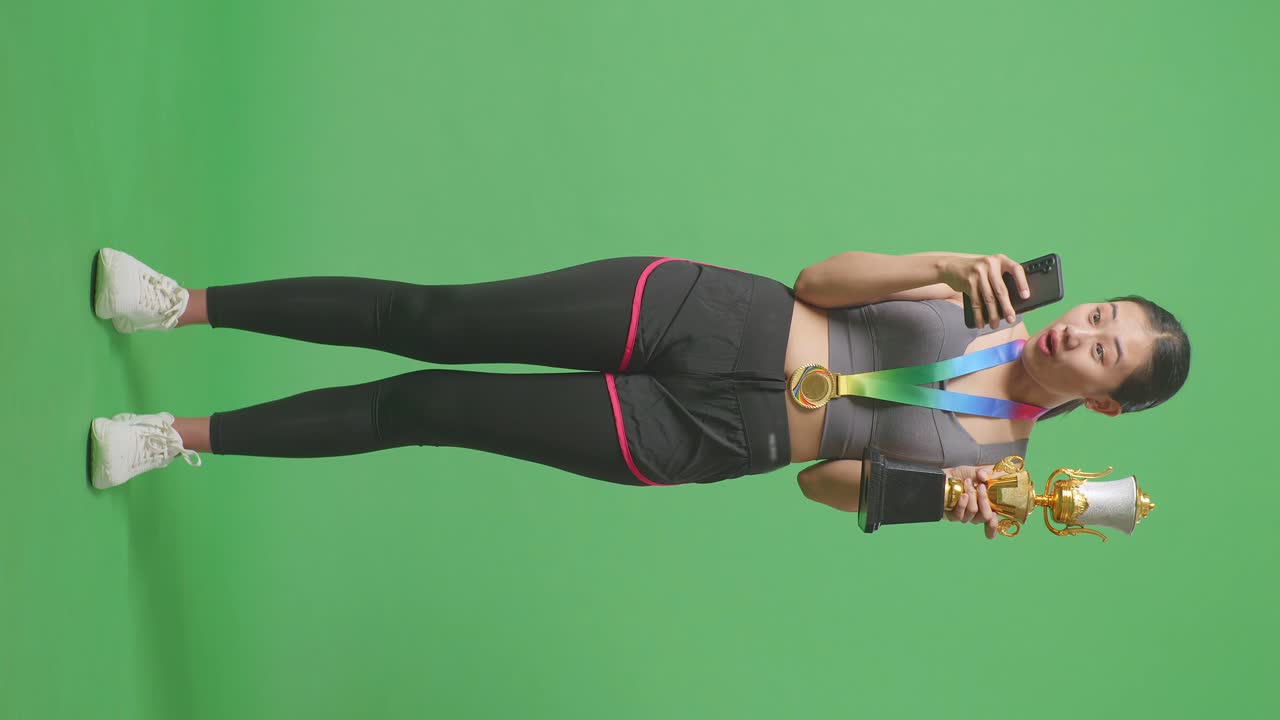 Full Body Of Asian Woman With A Gold Medal And Trophy Looking At Smartphone Then Saying Wow On Green Screen Background In The Studio